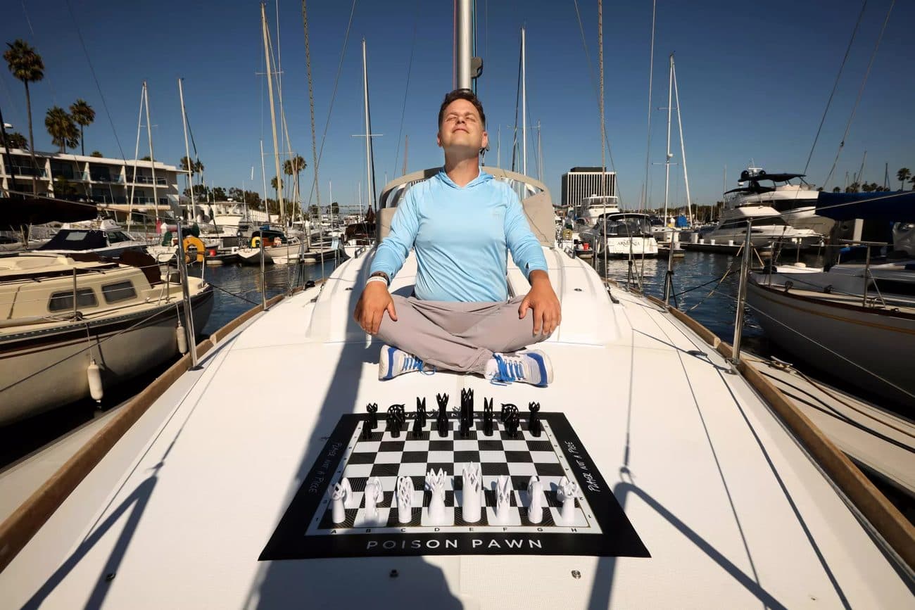 Seth Makowsky sits cross-legged, meditating on the deck of a sailboat with a Poison Pawn board in front of him.